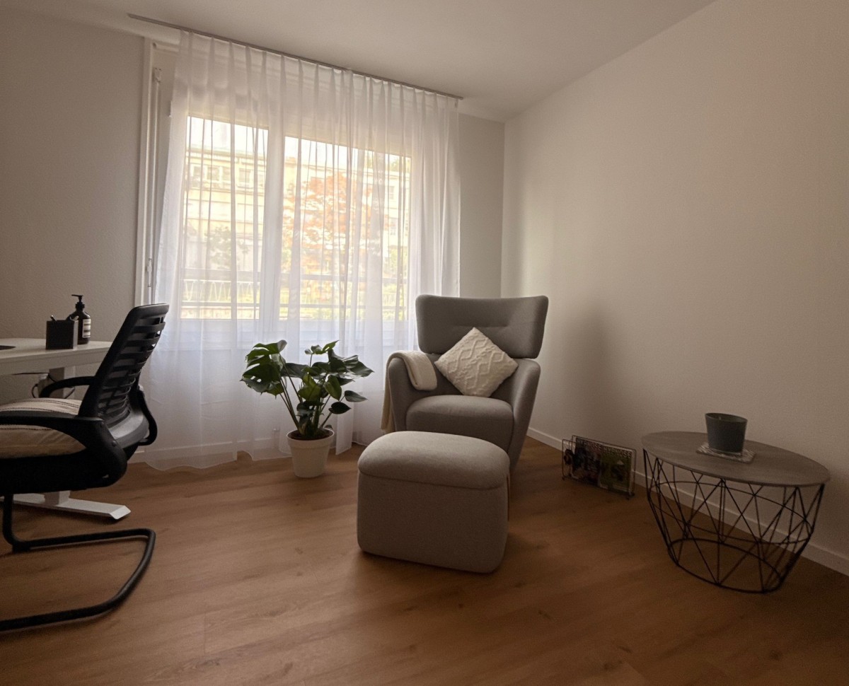 Salle de consultation du cabinet de Lausanne avec un fauteuil, un bureau et une plante près de la fenêtre.